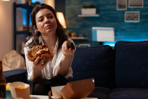 Caucasian female holding tasty burger in hands changing channels using remote watching comedy series