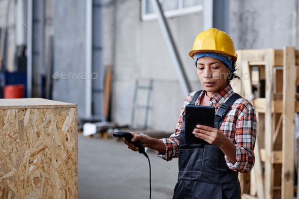 Young serious woman in coveralls and hardhat scanning barcode on huge box
