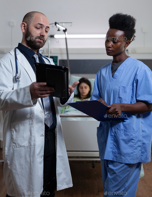 Doctor reading lab results from digital tablet to nurse holding clipboard with medical history at