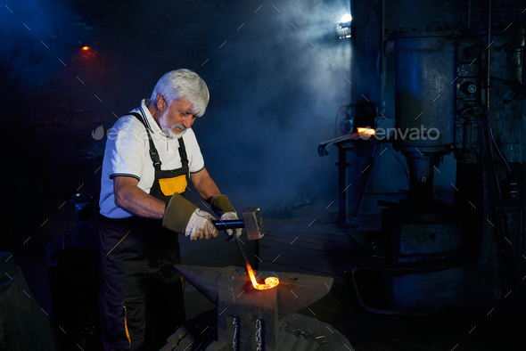Aged blacksmith forging steel on anvil with heavy hammer