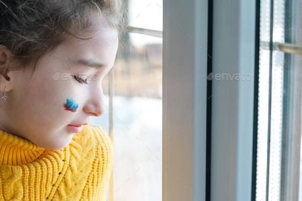 A sad child at the window with the flag of Russia, worries with tears in his eyes