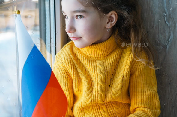 A sad child at the window with the flag of Russia, worries with tears in his eyes