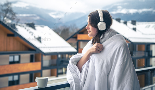 A young woman is listening to music wrapped in a blanket on the balcony.