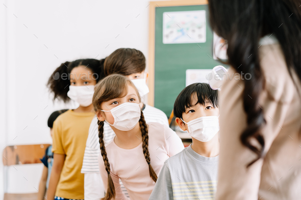 Multi-ethnic group of children with face mask stood in line let the teacher scan thermomete