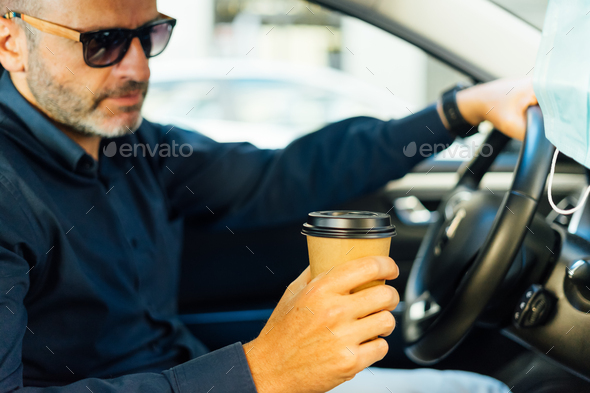 man drinking takeaway coffee inside the car with a protective mask hanging on the rearview mirror