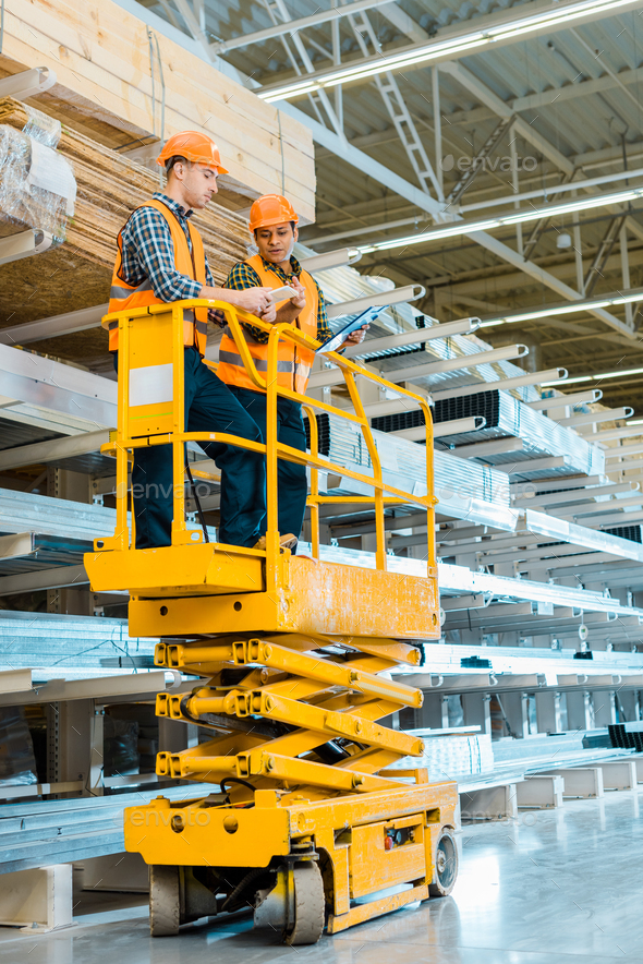 multicultural warehouse workers with digital tablet and clipboard standing on scissor lift