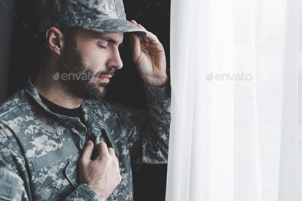 pensive bearded man in uniform holding hand on chest while standing by window with closed eyes