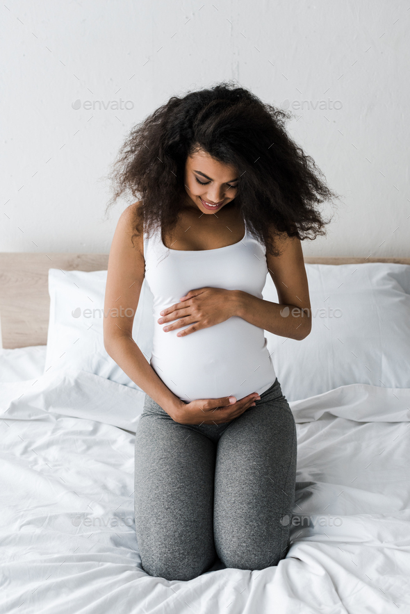 Curly African American Pregnant Woman Touching Belly While Sitting On