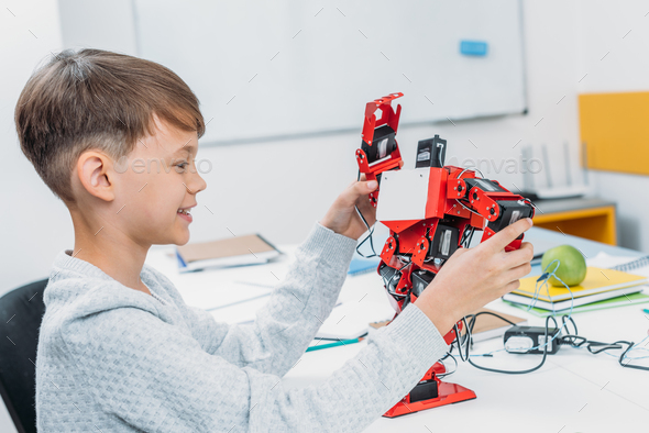 smiling schoolboy playing with red handmade robot in classroom