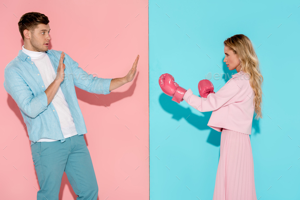 frightened man gesturing with hands near aggressive woman in pink boxing gloves on pink and blue