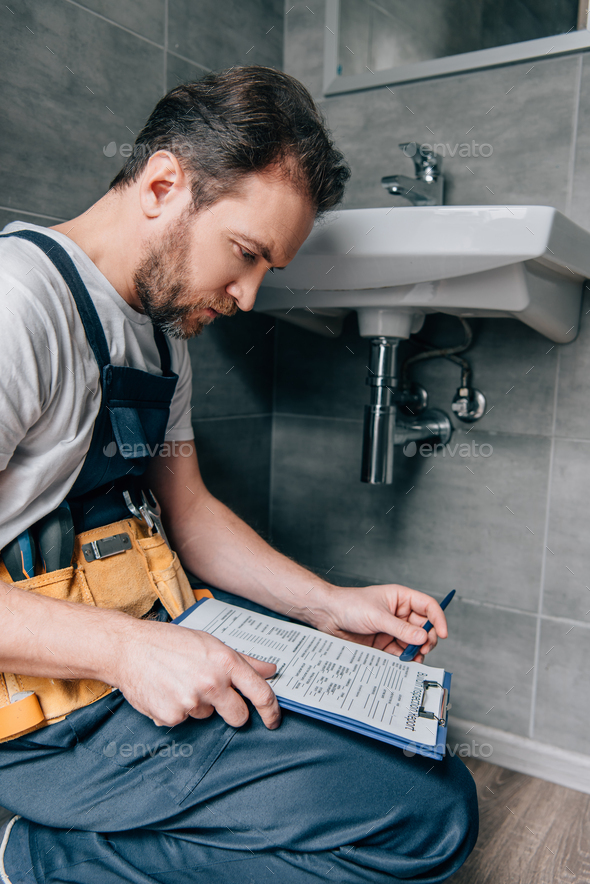 bearded male plumber with toolbelt writing in clipboard near broken sink in bathroom