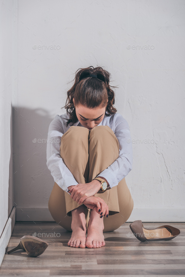 sad woman in white blouse and beige pants sitting on floor, hugging knees near wall at home,