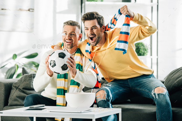 excited son and mature father with scarfs watching football and screaming at home