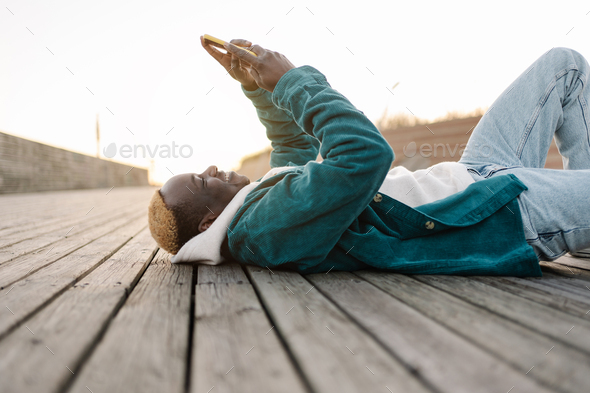 Happy black man holding smartphone using apps laying on wooden floor outdoors in the city - Stock Photo - Images