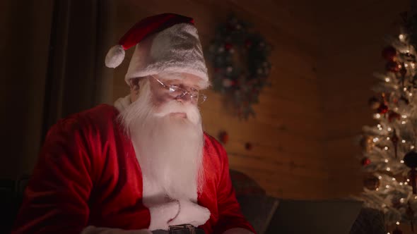 Santa Claus is Working on a Computer at Home with Glasses and a Beard Magical Christmas Lights on alt