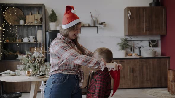 Mom Carefully Dress Red Santa Hat on the Head of Caucasian Son alt
