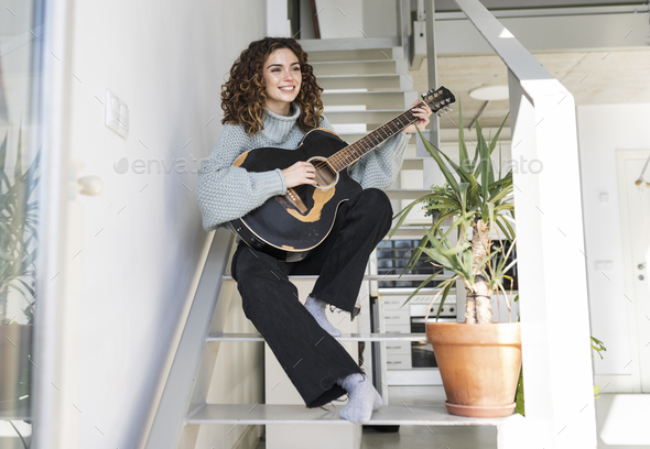 curly-haired young woman, on the stairs of the house, playing the guitar facing the sun