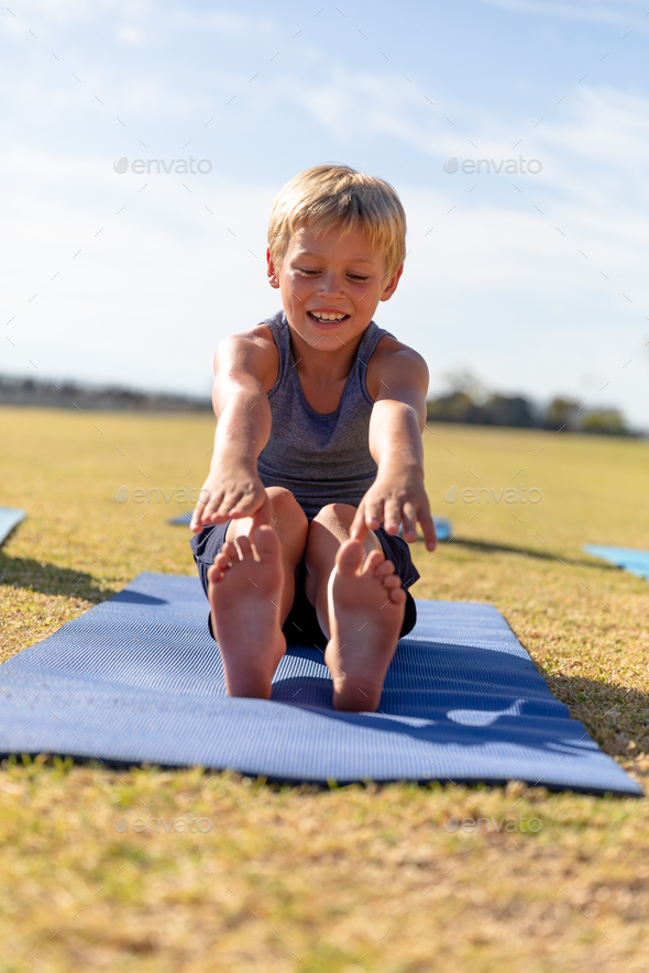 Full length of smiling caucasian elementary schoolboy touching toes while exercising on yoga mat