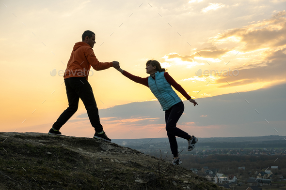 Man and woman hikers helping each other to climb stone at sunset in mountains. Couple climbing on
