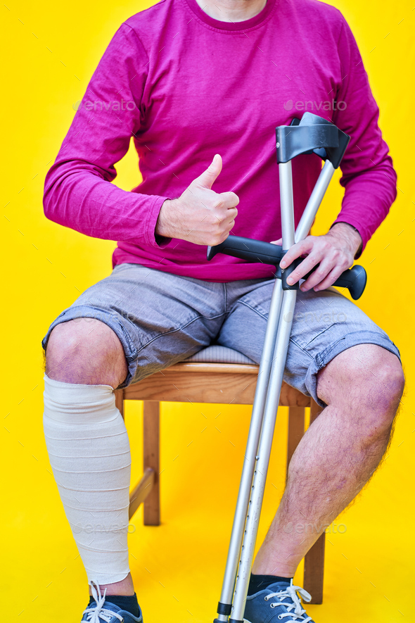 Man with crutches, jeans and purple shirt sitting on a chair with his thumb up on yellow background - Stock Photo - Images