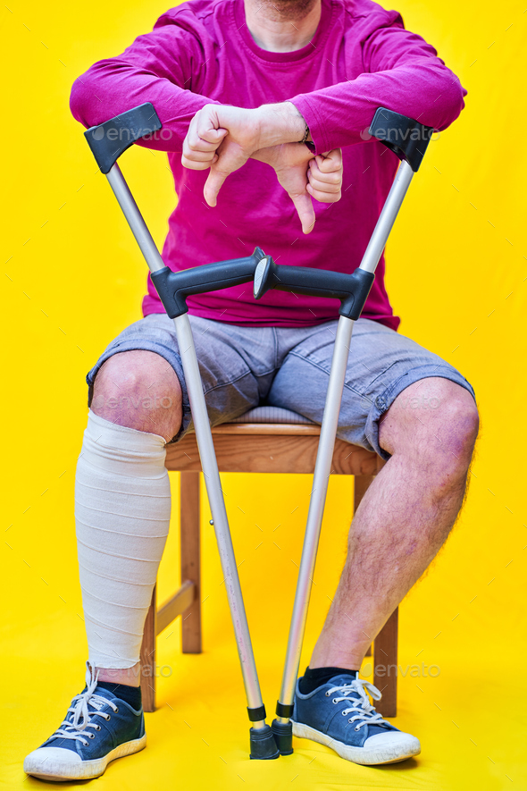 Man with crutches, sitting on a chair with his thumb down, on yellow background - Stock Photo - Images