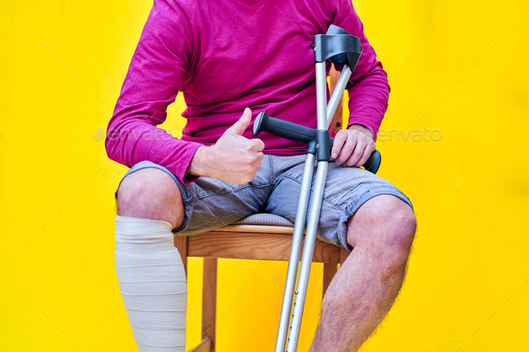 Man with crutches, sitting on a chair with his thumb up, on yellow background - Stock Photo - Images