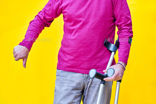 Man on crutches, jeans and purple t-shirt with his thumb down, on yellow background - Stock Photo - Images