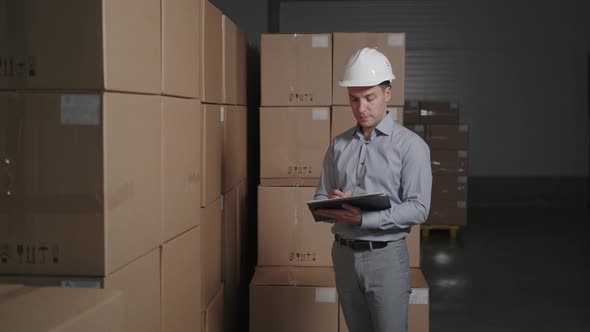 A Man Works in a Warehouse with Cardboard Boxes, Makes an Inventory