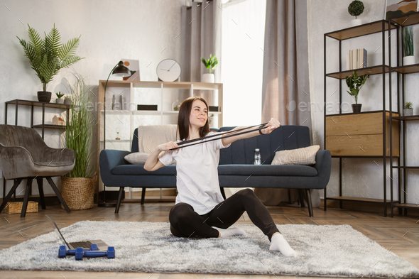 Attractive sporty woman dressed in sportsclothes, holds resistance band, having workout at home