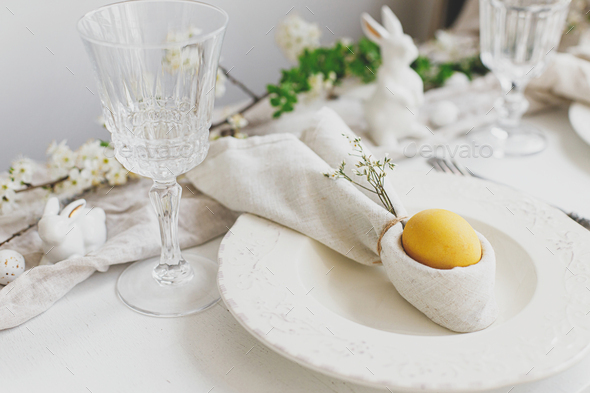 Stylish elegant Easter brunch table setting. Easter egg in bunny napkin, plate, cutlery, flowers