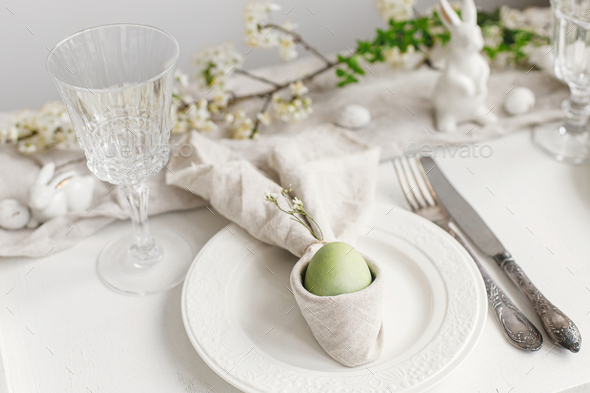 Stylish elegant Easter brunch table setting. Easter egg in bunny napkin, plate, cutlery, flowers