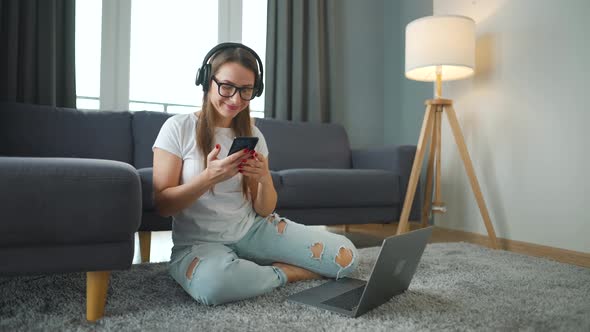 Casually Dressed Woman with Headphones is Sitting on Carpet with Laptop and Smartphone and Working alt