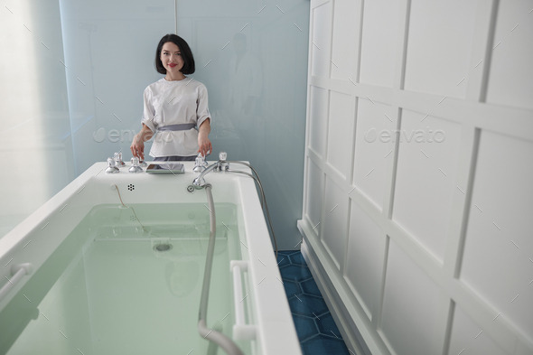 Smiling assistant prepares hydro massage bath full of clear water in spa salon