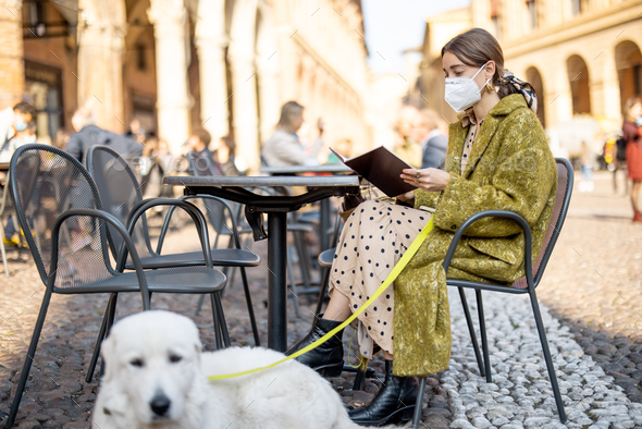 Woman in medical mask sitting with her dog at outdoor cafe in old italian city