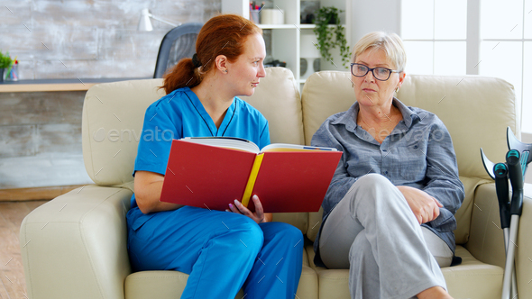 Caucasian female assistant in nursing home reading a book to an elderly retired woman