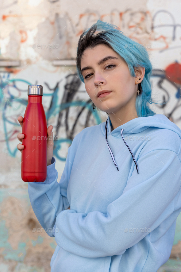 Blue haired Teenage girl in blue hoodie staying near graffiti wall with red water  bottle Stock Photo by katrinshine