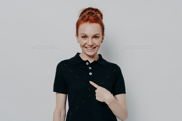 Young cheerful caucasian red haired woman pointing at herself while standing isolated over grey wall