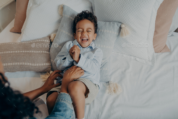 Adorable little son lying on pillows in bed and laughing while mom tickling him