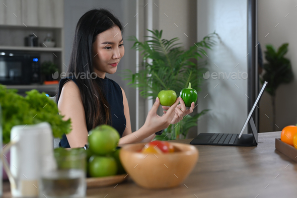 Young housewife holding green apple and bell pepper while having video call on digital tablet.