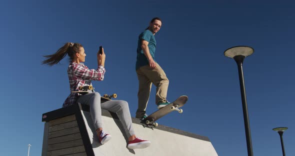 Happy caucasian woman taking photo of her male friend skateboarding alt