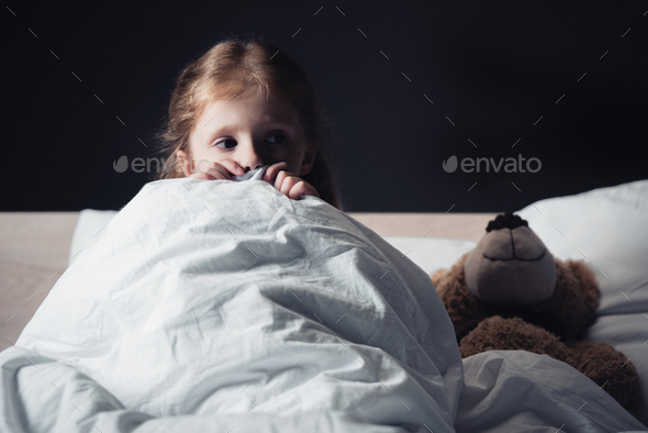 scared kid looking away while sitting under blanket near teddy bear isolated on black