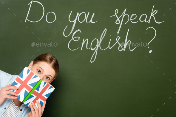 pretty girl with obscure face holding book with uk flag near chalkboard with do you speak English