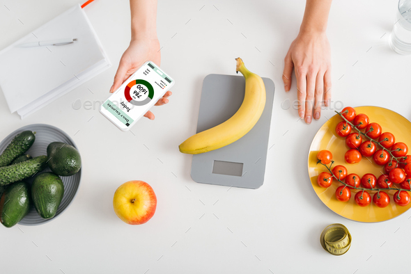 Top view of girl holding smartphone with calorie counting app while weighing banana on kitchen table