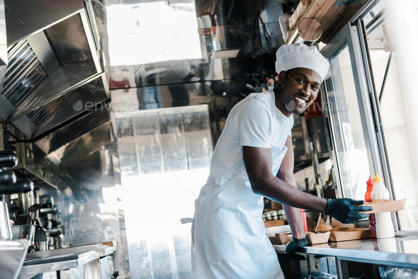 selective focus of cheerful african american chef smiling while holding carton plate in food truck