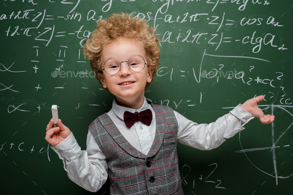 smiling kid in suit and bow tie holding chalk and pointing with hand at chalkboard with mathematical