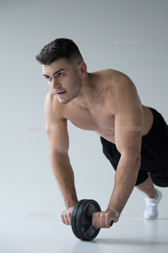 sexy muscular bodybuilder with bare torso exercising with ab wheel on grey background