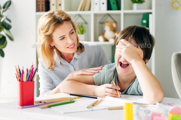 upset kid with dyslexia obscuring face and crying, smiling child psychologist looking at him