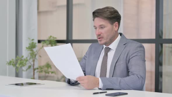 Successful Middle Aged Man Reading Documents while using Laptop alt