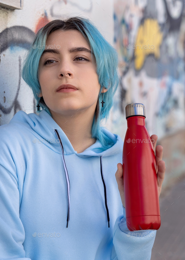 Blue haired Teenage girl in blue hoodie staying near graffiti wall with red water  bottle Stock Photo by katrinshine