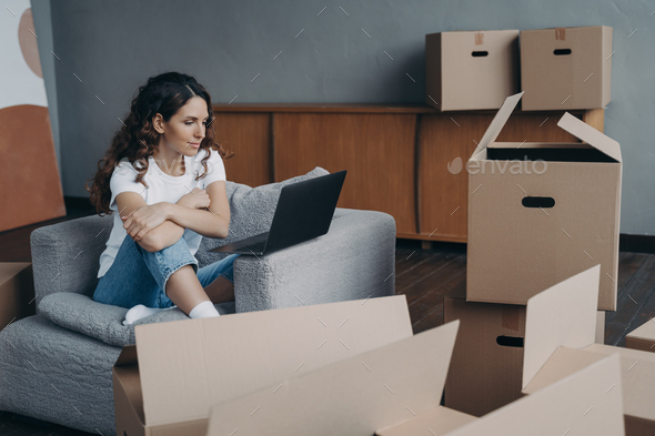 Happy young european woman selling apartment through internet. Girl among boxes is using laptop.